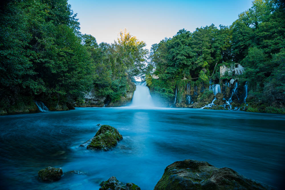 Langzeitbelichtung von einem Wasserfall und dem daraus fließenden Fluss. Das Wasser ist verschwommen und zieht weich durch die grüne Landschaft. Krka Nationalpark Kroatien
