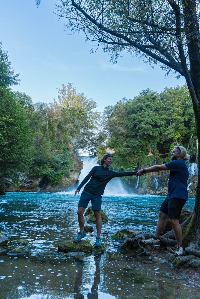 Melanie und Julian stehen vor einem Wasserfall und schauen sich an. Melanie steht auf einem Stein im Wasser und Julian versucht sie zurück zu ziehen. Krka Nationalpark Kroatien