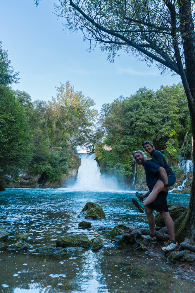 Melanie und Julian stehen vor einem Wasserfall und schauen sich an. Melanie sitzt Huckepack auf Julians Rücken. Beide lachen und schauen in die Kamera. Krka Nationalpark Kroatien