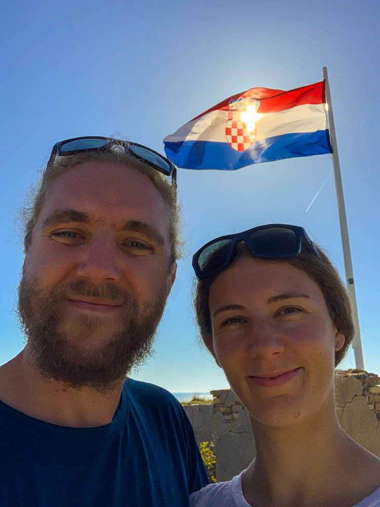 Selfie von Melanie und Julian an einem Lost Place in Kroatien mit der kroatischen Flagge im Hintergrund. Der Himmel ist kräftig blau, die Sonne scheint durch die Flagge