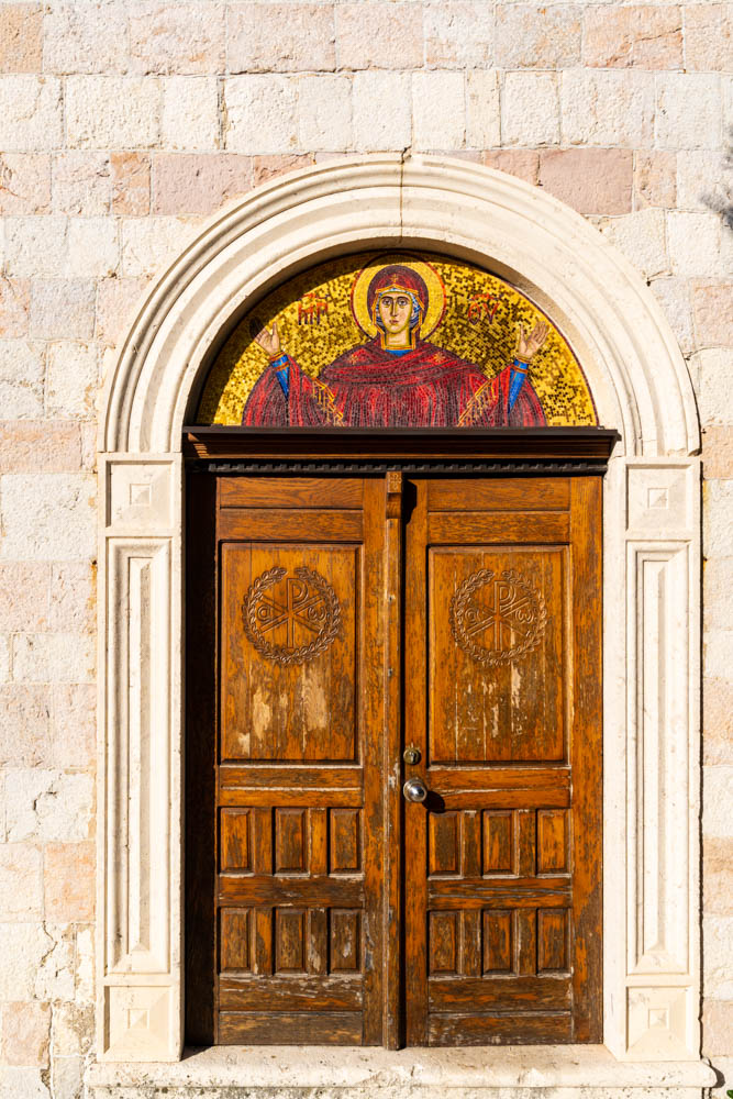 Holztür einer Kirche mit Rundbogen in Budva. Über der Tür ist eine Zeichnung eines Heiligen, der die Hände nach oben streckt.