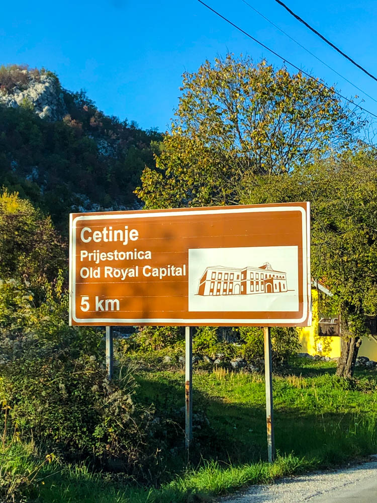 Schild am Straßenrand "Cetinje Old Royal Capital - 5 km". Dahinter sind einige Bäume zu sehen sowie ein kleiner Hügel. Der Himmel ist kräftig blau.