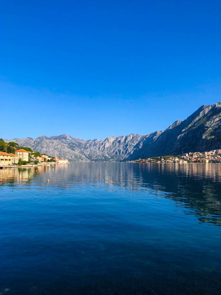 Kotor und Kotor Bucht "Fjord" in Montenegro. Der Himmel ist kräftig blau. Rechts und links sind Häuser zu sehen, im mittleren Teil des Bildes ist Wasser zu sehen. Alle Häuser sind umgeben von Bergen. Roadtrip