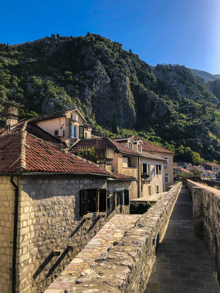Stadtmauer Kotor mit ein paar Steinhäusern links vom Bild. Dahinter ist eine grüne Hügellandschaft zu sehen. Der Himmel ist kräftig blau. Roadtrip Montenegro