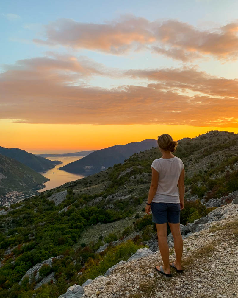 Melanie steht neben einer Klippe und blickt hinab in die Kotor Bucht in Montenegro. Der Himmel ist vom Sonnenuntergang orange rot verfärbt und glüht richtig. Roadtrip Montenegro