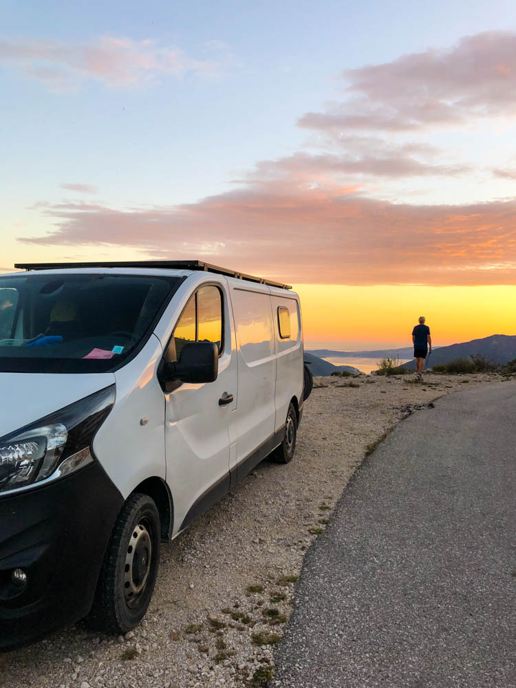 Van Vivaldi steht am Seitenrand einer Straße oberhalb der Kotor Bucht in Montenegro. Julian steht etwas weiter vorne und blickt ins Tal. Der Sonnenuntergang hat den Himmel orange rot verfärbt. Diese Färbung spiegelt sich im Camper. Roadtrip