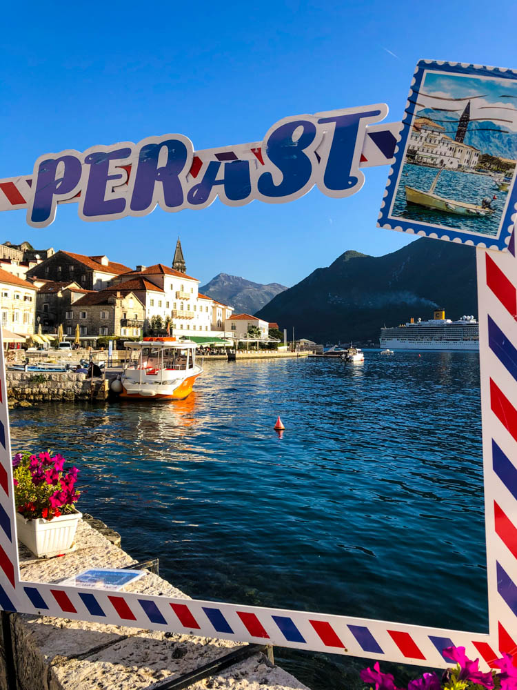 Das Bild sieht aus wie eine Postkarte. Ein Rahmen, über welchem Perast steht und rechts oben eine Briefmarke klebt, steht vor Perast mit der Stadt im Fokus. Kräftig blauer Himmel, das Meer sowie die Stadt Perast ergeben ein idyllisches Bild. Roadtrip Montenegro