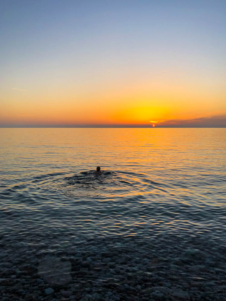Die Sonne geht über dem Meer unter und verfärbt den Himmel sowie das Wasser orange rot. Julian schwimmt im Meer und beobachtet den Sonnenuntergang dabei. Aufnahme bei Sveti Stefan - Roadtrip Montenegro