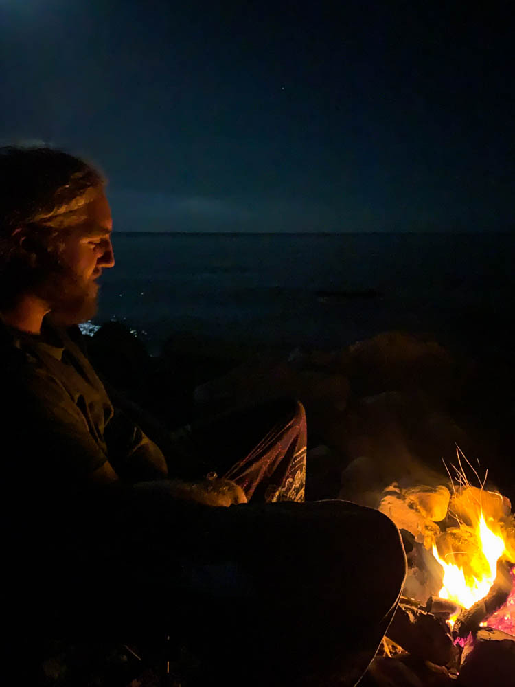 Julian sitzt am Lagerfeuer direkt am Meer und blickt in die Flammen. Roadtrip Montenegro Vanlife