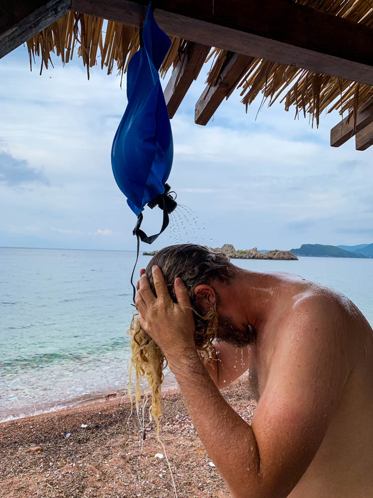 Julian duscht sich mit einem Wassersack nach dem Schwimmen im Mittelmeer. Er steht dabei unter einer Hütte, welche mit Stroh bedeckt ist. Roadtrip Vanlife Montenegro