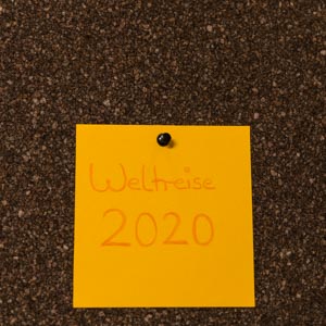 Ein gelber Zettel mit der Aufschrift Weltreise 2020 ist an eine dunkle Korkpinnwand gepinnt.