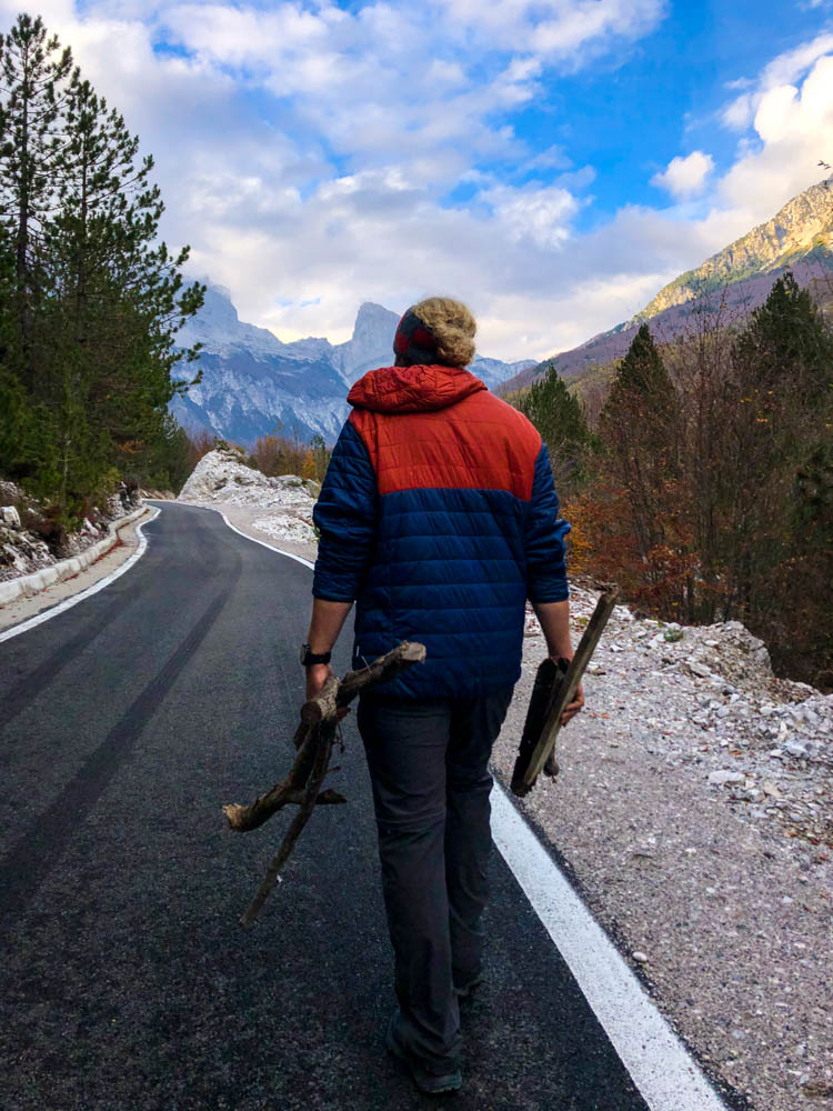 Julian läuft auf einer geteerten Straße in Albanien bei Theth spazieren und sammelt gerade Feuerholz. Vor ihm ragt eine schöne Bergkulisse auf.