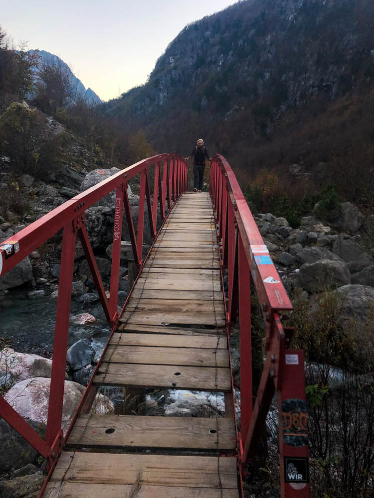 Julian läuft über eine Brücke, welche über eine Schlucht führt. Es fehlen einige Holzbretter der Brücke, weshalb Julian sich am roten Geländer festhält. Albanische Gebirgslandschaft