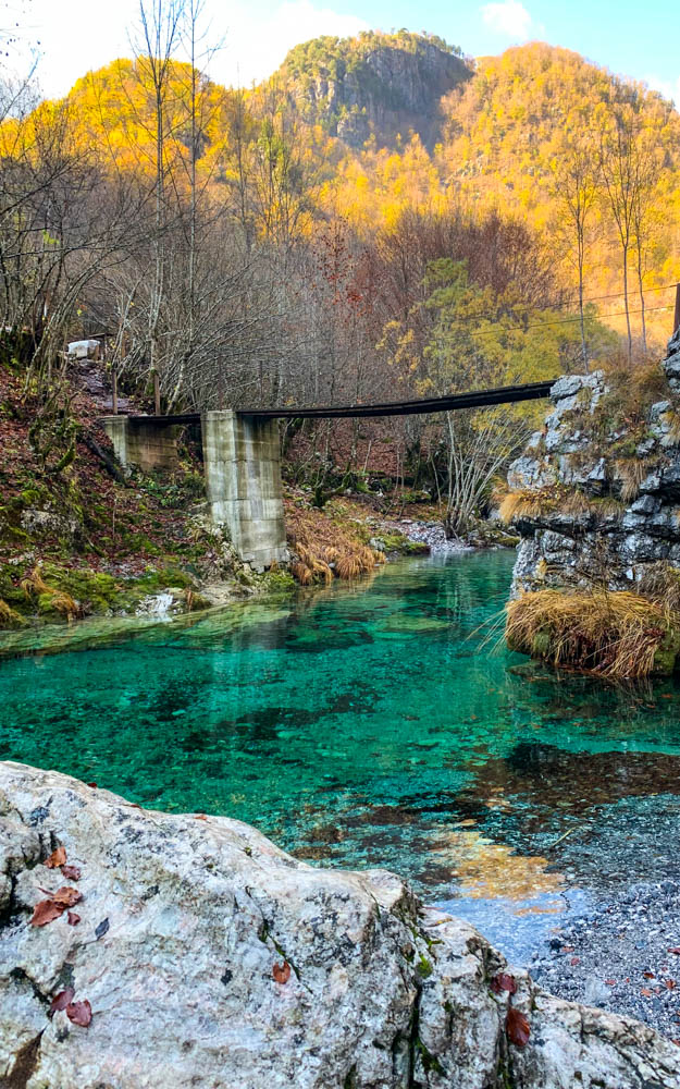 Landschaftsaufnahme Albanien. Eine Brücke führt über einen türkisfarbenen Fluss. Dahinter ist ein herbstlich verfärbter Wald zu sehen.