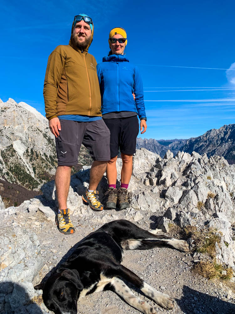 Gipfelaufnahme von Melanie und Julian am Valbona Pass im Nationalpark Theth in Albanien. Vor ihnen liegt ein Straßenhund, welcher ebenfalls den Pass hochgelaufen ist. Gebirgslandschaft mit kräftig blauem Himmel