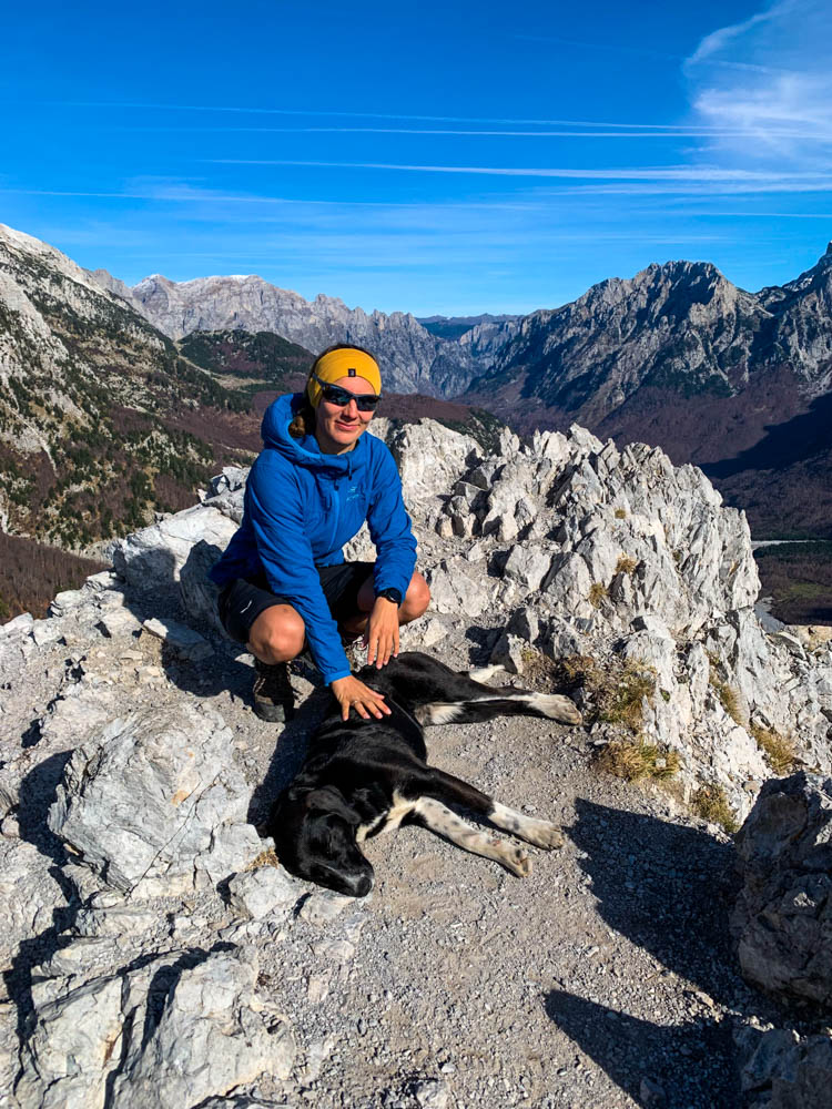 Melanie am Gipfel des Valbona Pass im Nationalpark Theth in Albanien. Sie grinst in die Kamera und streichelt dabei einen Straßenhund. Hinter ihr ist das Valbona Tal mit einigen Bergen zu sehen, der Himmel ist kräftig blau.