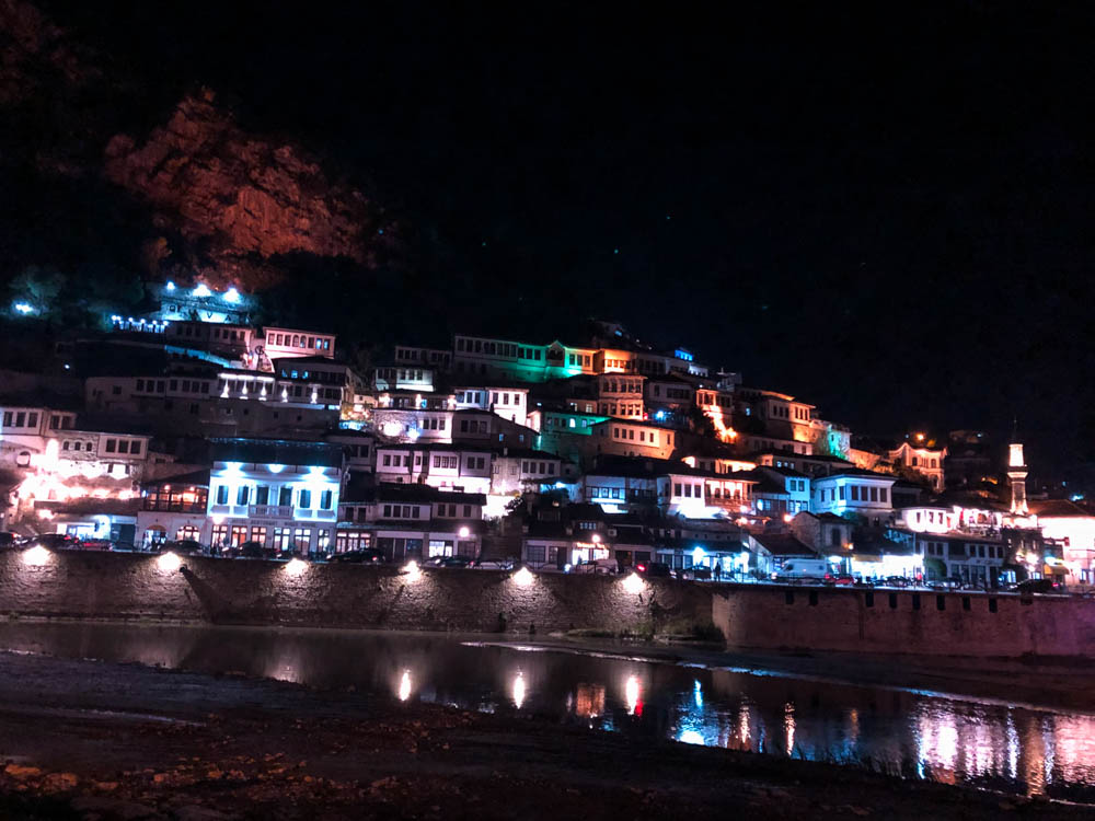 Berat, die Stadt der tausend Fenster, bei Nacht. Die Häuser sind in unterschiedlichen Farben beleuchtet und spiegeln sich im Fluss wieder. Albanien - ein Land voller Gegensätze