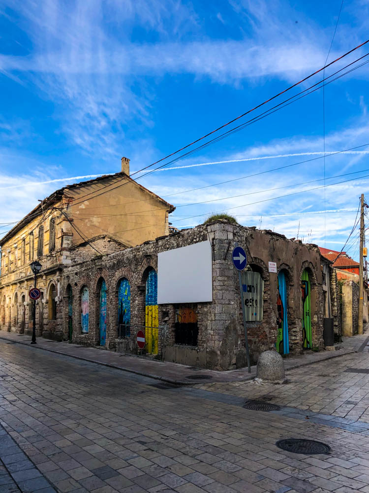 Streetart in Shkodra - mehrere Türen und Fenster einer Häuserfassade sind bunt mit verschiedenen Motiven bemalt. Der Himmel ist schön blau mit ein paar Schleierwolken. Albanien - ein Land voller Gegensätze