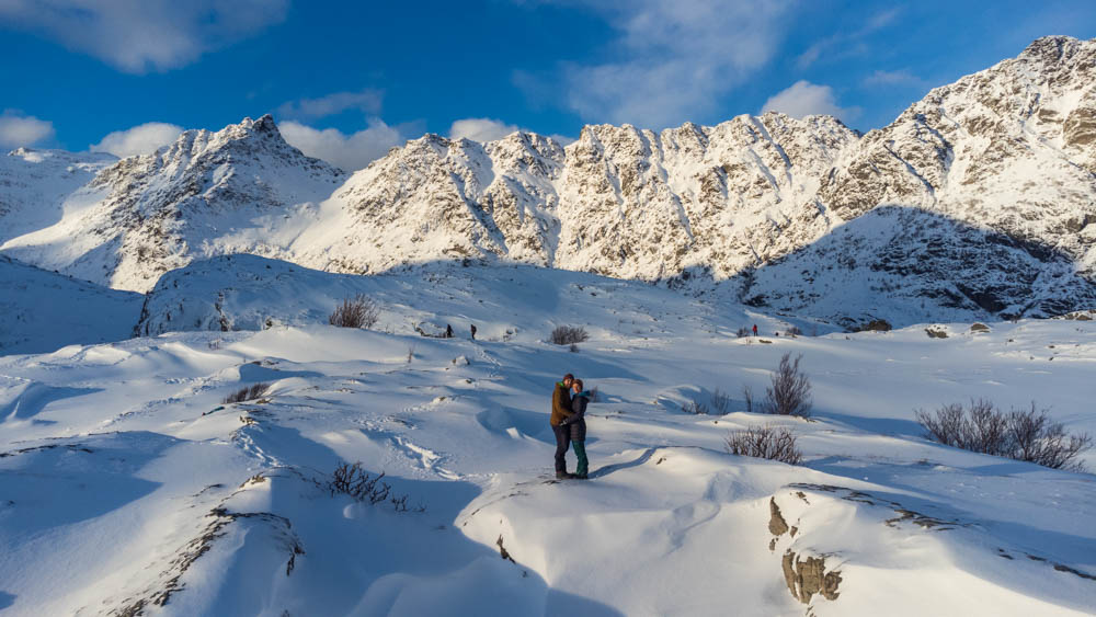 Aussichtspunkt am Ende der Lofoten bei Ä. Verschneite Winterlandschaft mit toller Bergkulisse. Melanie und Julian stehen mitten drin, halten sich gegenseitig fest und blicken zur Drohne, welche etwas vor ihnen schwebt.
