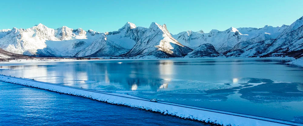 Van Vivaldi fährt über eine Brücke am Grunnførfjord auf den Lofoten. Verschneite Winterlandschaft, Berge spiegeln sich im Wasser und weißer Van fährt über eine vom Schnee weiße Straße. Der Himmel ist kräftig blau.