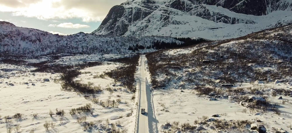 Camper Van Vivaldi fährt durch winterliche Landschaft auf zugeschneiter Straße. Kerzengerade Straße auf dem Weg nach Nusfjord auf den Lofoten, Norwegen