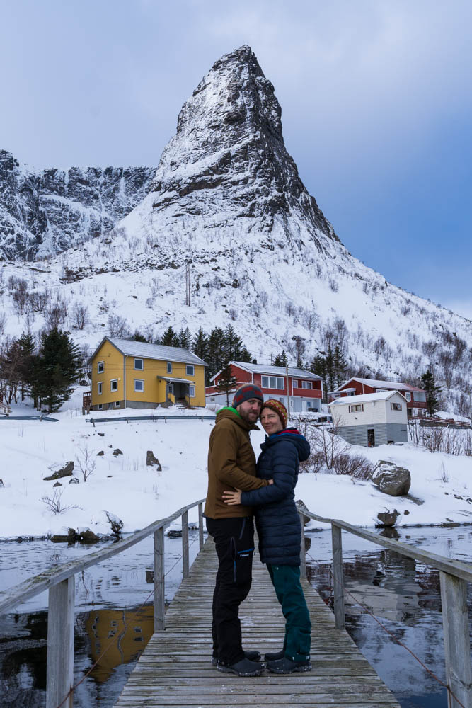 Melanie und Julian stehen eng zusammen auf einer Brücke in Reine, hinter ihnen ist der Berg Horn zu sehen. Winterliche Landschaft auf den Lofoten in Norwegen