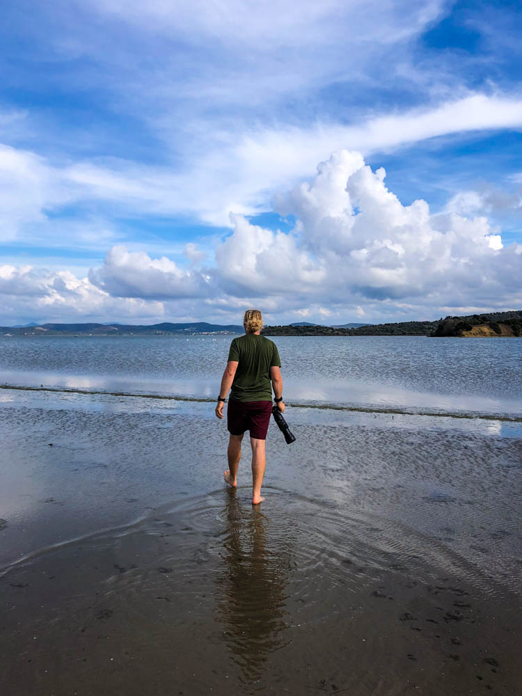 Julian mit Teleobjektiv und Kamera an Lagune. Er läuft barfuß am Strand entlang und vor ihm sind klein ein paar Flamingos im Wasser zu erkennen