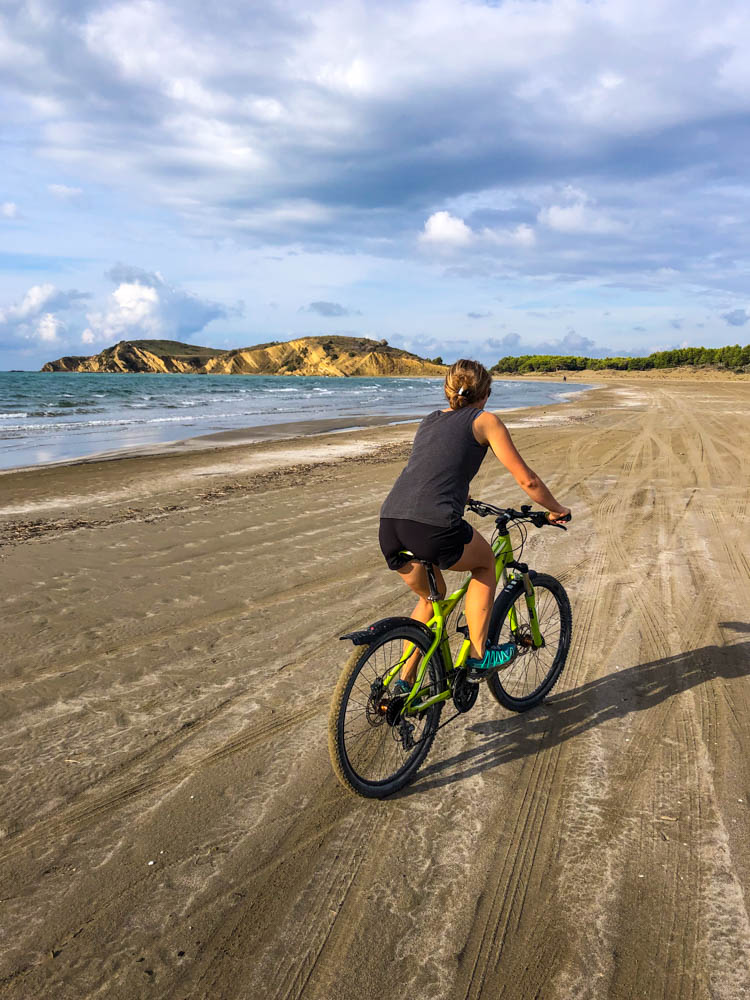 Melanie fährt mit dem Fahrrad über einen Sandstrand. Links ist das Meer zu erkennen, vor ihr liegen ein paar kleinere Hügel. Südalbanien