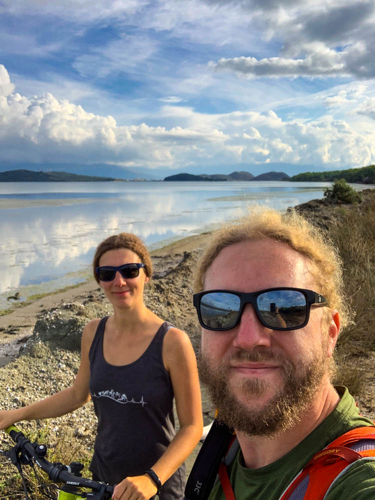 Selfie von Melanie und Julian bei Narta Lagune Albanien. Hinter ihnen ist das Wasser der Lagune zu sehen. Die Wolken spiegeln sich im Wasser.