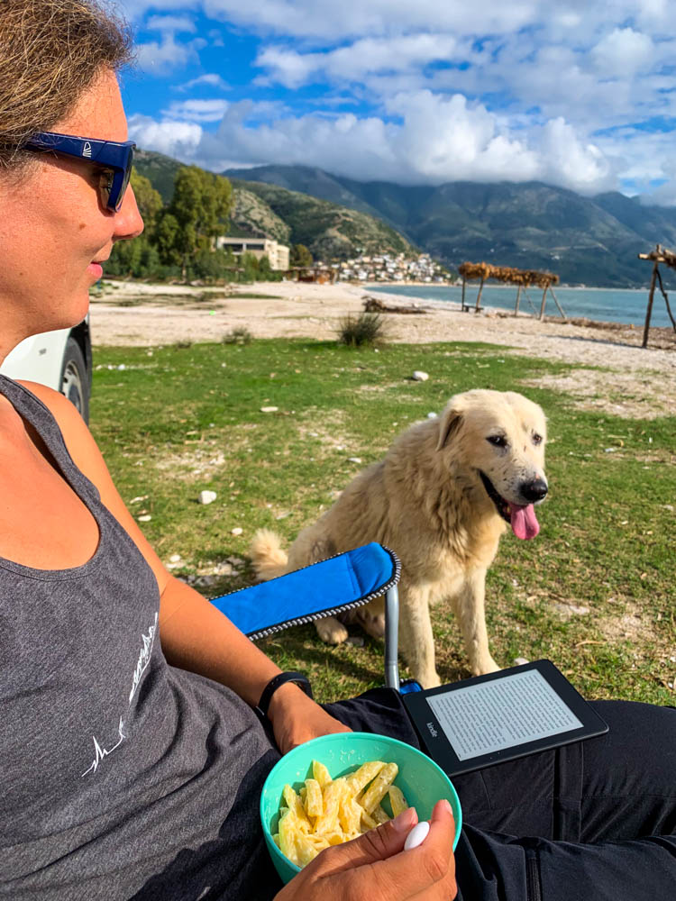 Abendessen in Südalbanien - Melanie sitzt auf ihrem Campingstuhl und wird von einem Straßenhund beobachtet. Herrliche Landschaft im Hintergrund mit Hügeln, Strand und grünen Tönen