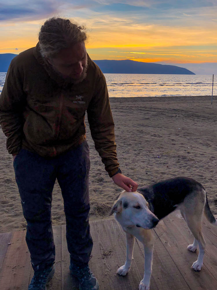 Julian streichelt ein Straßenhund. Hinter ihm geht die Sonne über dem Meer unter. Sonnenuntergang in Südalbanien