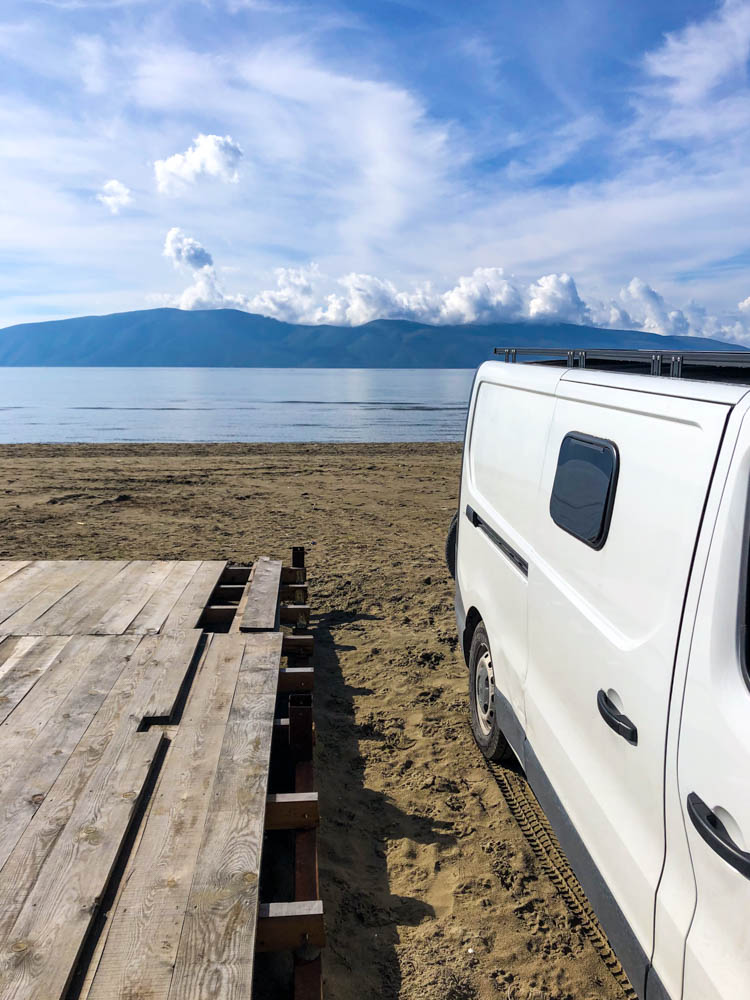Van Vivaldi steht neben einer Holzplattform im Sand eines Strandes in Albanien bei Vlora. Hinter ihm ist das Meer zu sehen. Der Himmel ist leicht bewölkt.