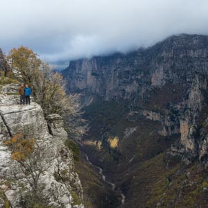 Vikos Schlucht in Griechenland im Herbst. Ein Fluss schlängelt sich durch die Schlucht, rechts und links ragen die Felswände in die Höhe. Melanie und Julian stehen links auf einem Felsen und blicken in die Kamera.