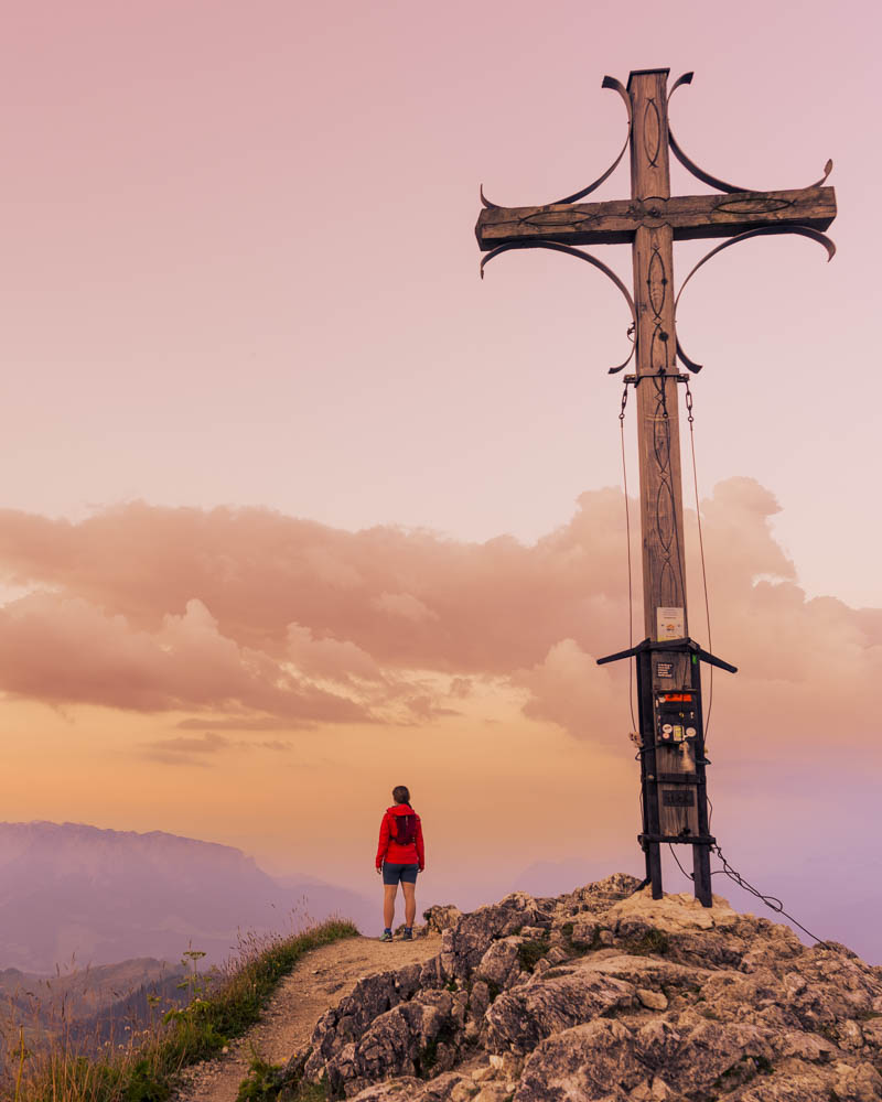 Melanie steht neben dem großen Gipfelkreuz des Geigelstein und blickt auf das Kaisergebirge. Sonnenaufgang in den Chiemgauer Alpen