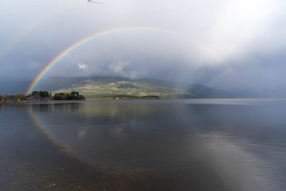 Regenbogen spiegelt sich in einem See in Ioannina in Griechenland. Über dem Regenbogen ist noch recht schwach ein zweiter Regenbogen zu erkennen.