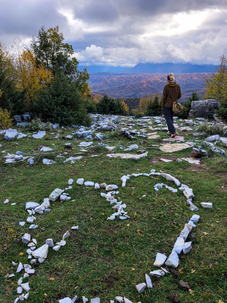 Julian blickt in die Ferne ins Pindos Gebirge und steht dabei im Stone Forest in Griechenland. Im Vordergrund ist ein mit Steinen gelegtes Herz zu sehen.