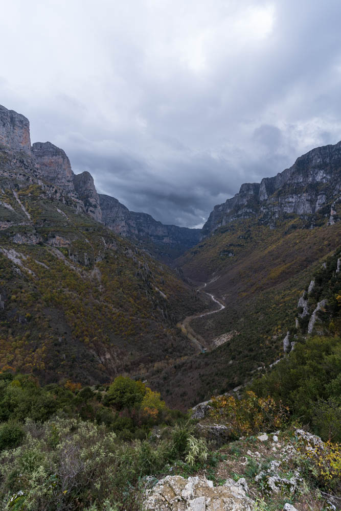 Vikos Schlucht in Griechenland im Herbst. Ein Fluss schlängelt sich durch die Schlucht, rechts und links ragen die Felswände in die Höhe. Düstere Wolken hängen über der Schlucht