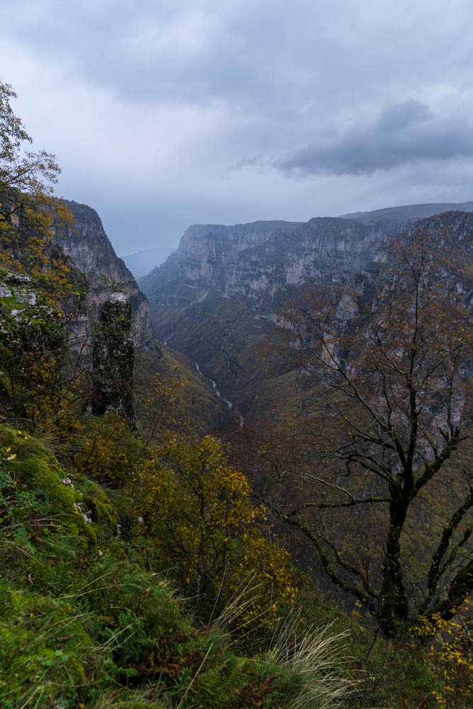 Vikos Schlucht in Griechenland im Herbst. Ein Fluss schlängelt sich durch die Schlucht, rechts und links ragen die Felswände in die Höhe. Düstere Wolken hängen über der Schlucht