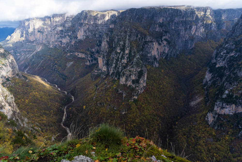 Vikos Schlucht in Griechenland im Herbst. Ein Fluss schlängelt sich durch die Schlucht, rechts und links ragen die Felswände in die Höhe. Eine weitere Schlucht ist zu sehen und die Sonne kämpft sich durch die Wolken