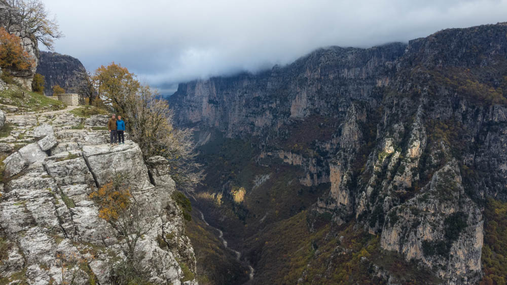 Vikos Schlucht in Griechenland im Herbst. Ein Fluss schlängelt sich durch die Schlucht, rechts und links ragen die Felswände in die Höhe. Melanie und Julian stehen links auf einem Felsen und blicken in die Kamera.