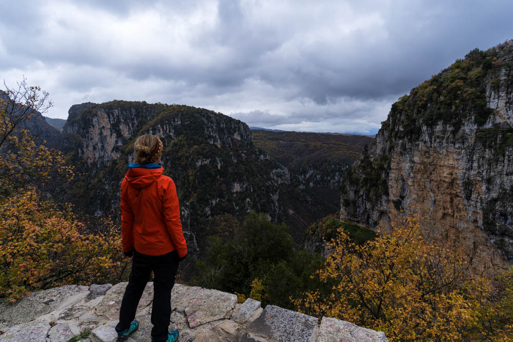 Melanie blickt in die Vikos Schlucht in Griechenland. Der Himmel ist bewölkt und gibt dem Bild eine düstere Stimmung. Die Herbstfarben der Natur sowie die orangene Jacke von Melanie erhellen das Bild farbenfroh.