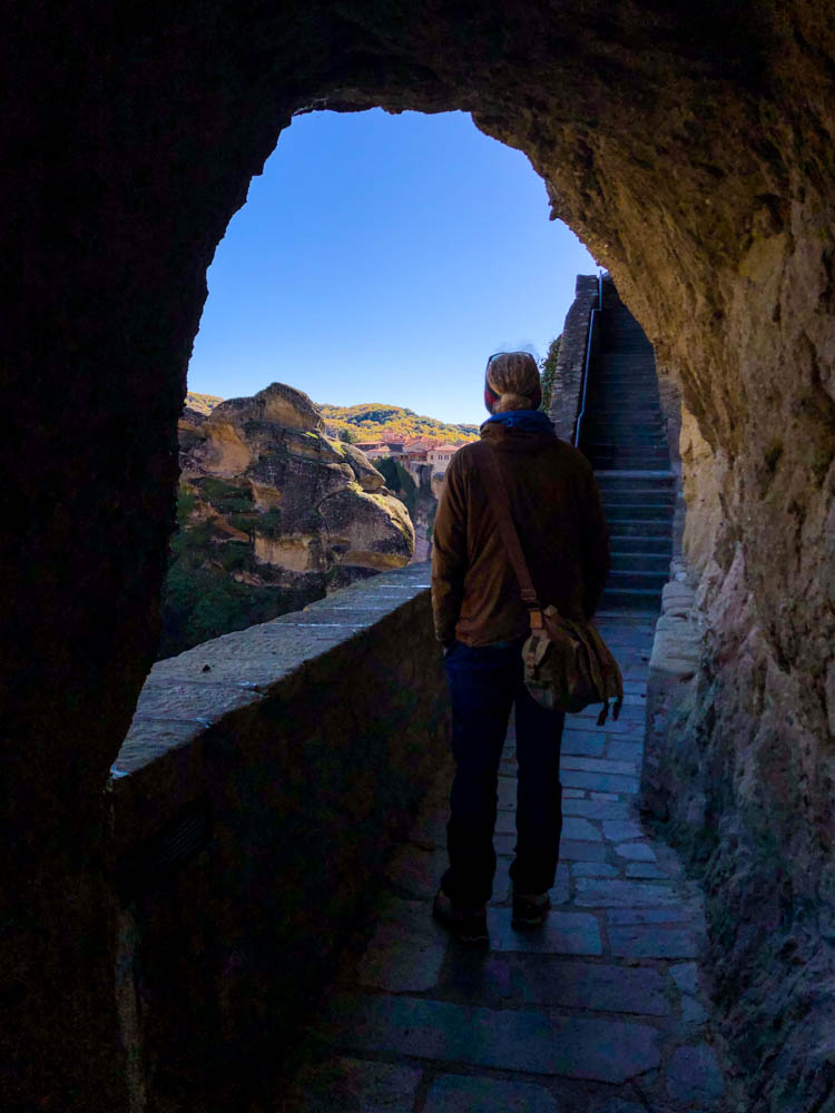 Julian läuft durch einen Steinbogen auf eine Treppe zu, welcher hinauf in ein Meteora Kloster führt. Vor ihm ist ein weiteres Kloster zu sehen. Griechenland im Herbst