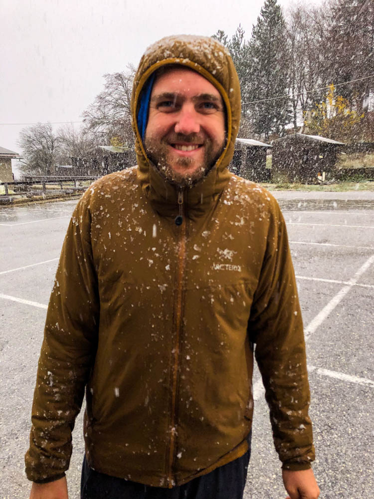 Julian steht auf einem griechischen Parkplatz und grinst in die Kamera. Es schneit kräftig.