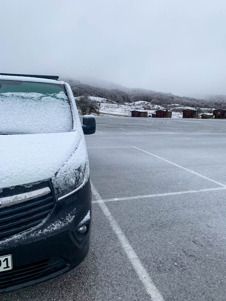 Van Vivaldi steht auf einem griechischen Parkplatz, welcher eine leichte Schicht Schnee trägt. Das Auto ist voller Schnee.