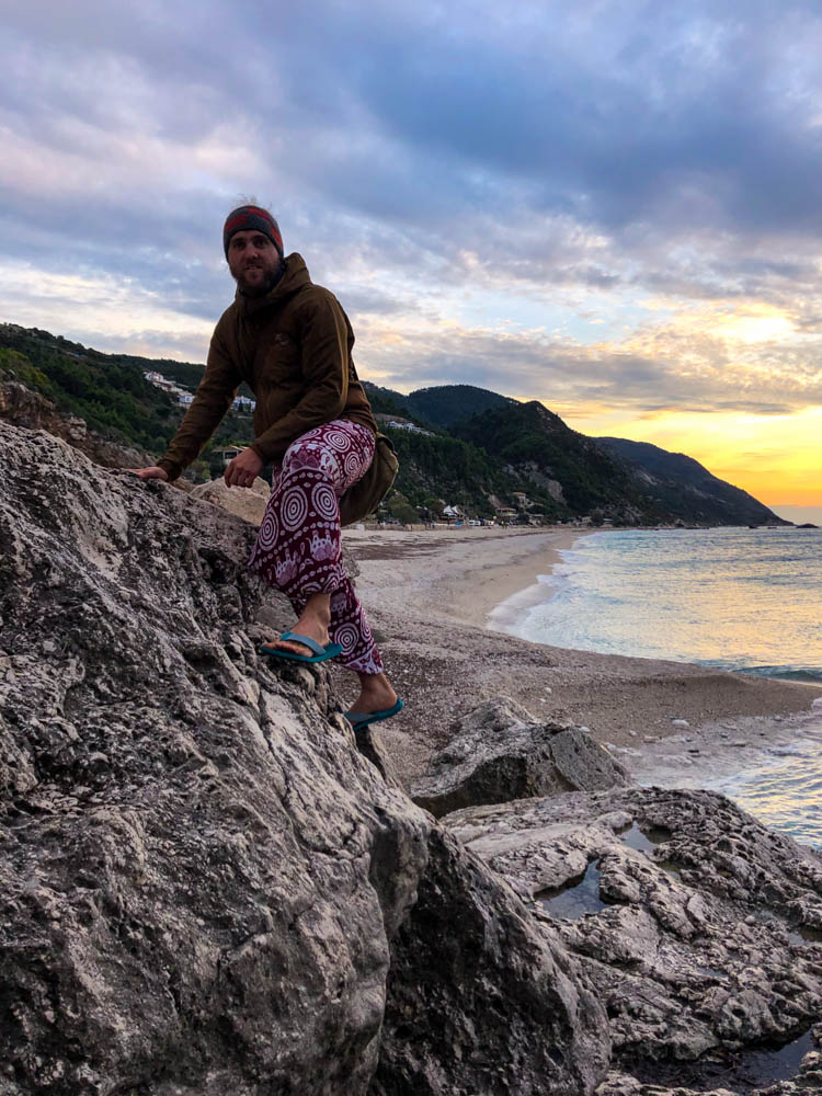 Julian klettert auf griechischen Insel Lefkada auf Felsblöcken herum. Hinter ihm sieht man den Strand sowie den rötlich verfärbten Himmel. Sonnenuntergang und zur Ruhe kommen