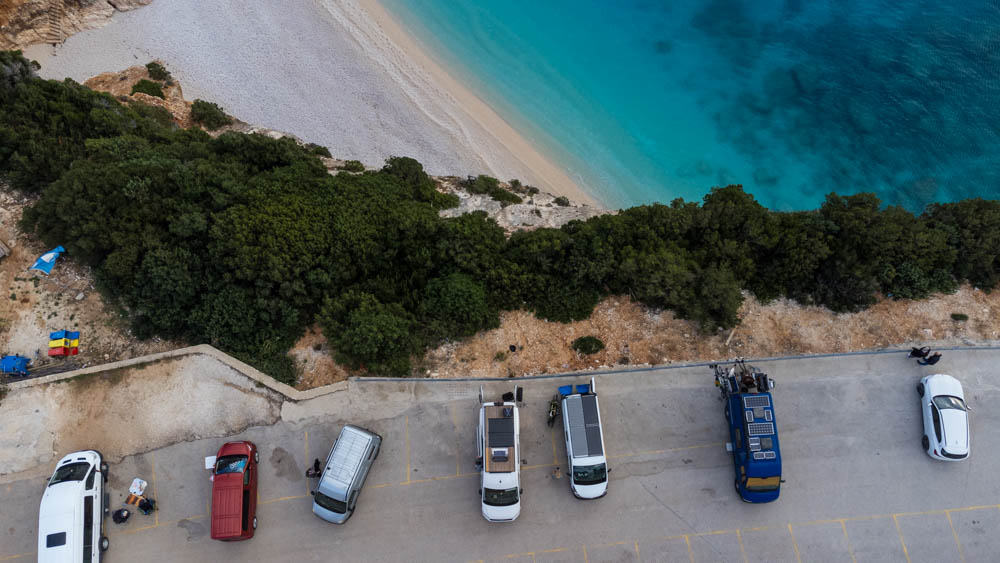 Vogelperspektive von Stellplatz in Griechenland auf Lefkada. Im unteren Teil sind ein paar Fahrzeuge auf einem Parkplatz zu sehen, im oberen Teil eine Strandbucht mit türkisfarbenem Wasser im Meer