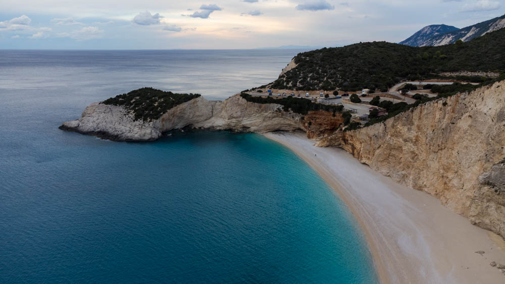 Drohnenaufnahme von Strandbucht auf Lefkada in Griechenland. Auf einer Anhöhe auf einem Parkplatz ist Van Vivaldi als kleiner Punkt leicht zu erkennen. Türkisfarbenes Wasser und teils bewaldete Hügellandschaft. Zur Ruhe kommen auf Lefkada im griechischen Paradies
