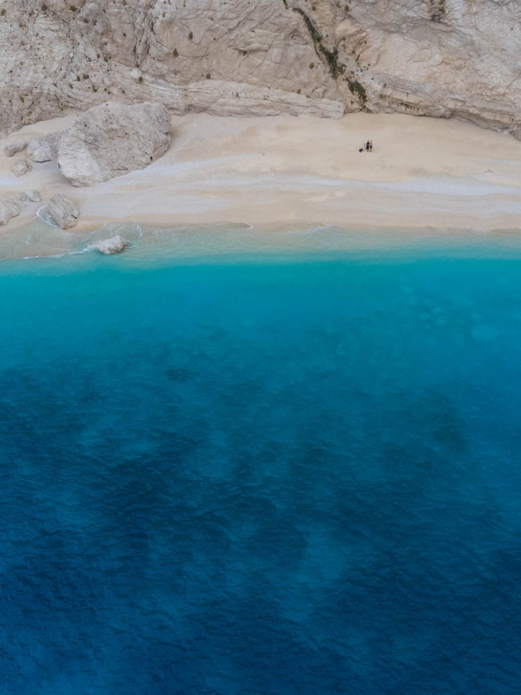 Luftaufnahme von Strandbucht auf Lefkada. Melanie und Julian sind als kleine Punkte an Strand zu erkennen, hinter ihnen ist eine Felswand. Im unteren Teil des Bildes ist das türkisfarbene Wasser zu sehen.
