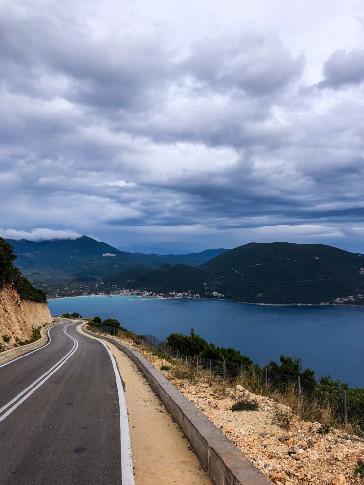 Kurvige Straße auf griechischer Insel Lefkada. Die Straße führt zu einer Bucht, der Himmel ist stark bewölkt.