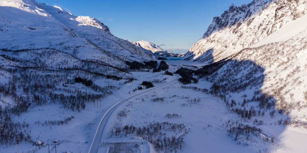 Luftaufnahme von Winterlandschaft Skandinavien. Es liegt Schnee auf der Straße, welche durch ein Bergtal führt. Der Himmel ist kräftig blau.